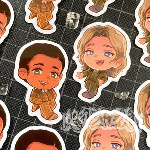Könnte beinhalten: Eine Sammlung von gestanzten Aufklebern mit Chibi-Figuren, die Vampirzähne haben. Eine Figur hat dunkle Haut und trägt einen beigen karierten Anzug, während eine andere blondes Haar hat und einen olivgrünen Anzug trägt. Beide Figuren haben ausdrucksstarke Gesichter.