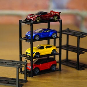 Peut inclure: Un support de stockage de voitures noir à plusieurs niveaux contient quatre voitures jouets. Les voitures sont rouges, jaunes, bleues et rouges. La voiture du haut est rouge avec des accents noirs. Le support est en plastique noir.