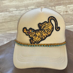 May include: A beige trucker hat with a brown and orange tiger embroidered on the front. The hat has a brown beaded trim around the brim.