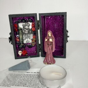 Puede incluir: Una peque&ntilde;a caja de madera con un interior de purpurina morada. La caja est&aacute; abierta y contiene una peque&ntilde;a estatua de Santa Muerte, una peque&ntilde;a calavera, una llave, un rosario rojo y negro y una peque&ntilde;a botella. La caja est&aacute; rodeada de un trozo de papel blanco con texto que dice "Oraci&oacute;n de apertura de camino a Santa Muerte".