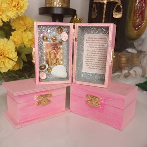 May include: Two small pink wooden boxes with gold latches. One box is open, revealing a silver glitter interior, a small image of a figure, white pearls, gold beads, a white seashell, and a printed text card dedicated to Aphrodite.