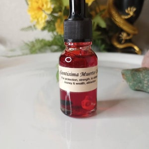 May include: A small glass bottle with a black dropper top filled with a red liquid. The bottle has a white label with the text "Santisima Muerte Oil" and a description of its uses.