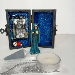 Puede incluir: Una peque&ntilde;a caja de madera con un interior de purpurina azul, que contiene una figura de Santa Muerte, una vela peque&ntilde;a y una peque&ntilde;a bolsa con una oraci&oacute;n a Santa Muerte. La caja tiene una cerradura y un peque&ntilde;o pestillo de metal.