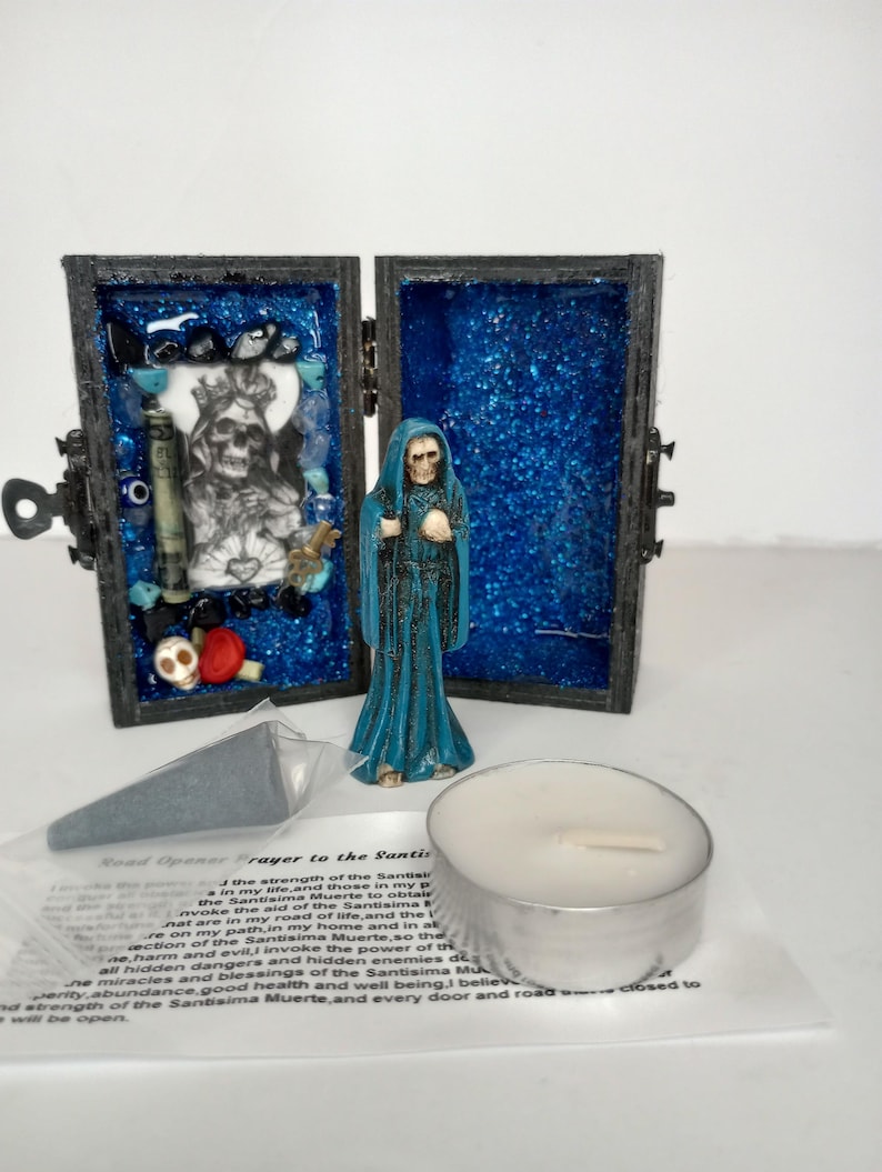 Puede incluir: Una peque&ntilde;a caja de madera con un interior de purpurina azul, que contiene una figura de Santa Muerte, una vela peque&ntilde;a y una peque&ntilde;a bolsa con una oraci&oacute;n a Santa Muerte. La caja tiene una cerradura y un peque&ntilde;o pestillo de metal.