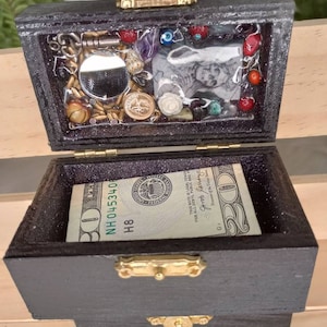 Puede incluir: Una peque&ntilde;a caja de madera con un cierre y bisagras doradas. La caja est&aacute; abierta y contiene un billete de 20 &euro;, un peque&ntilde;o espejo y varias baratijas, incluyendo una peque&ntilde;a foto, algunas cuentas y algunas monedas.