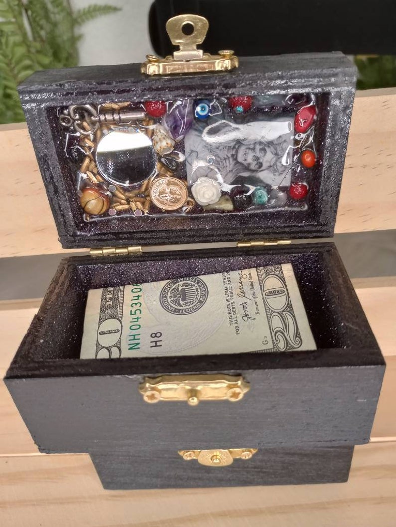 Puede incluir: Una peque&ntilde;a caja de madera con un cierre y bisagras doradas. La caja est&aacute; abierta y contiene un billete de 20 &euro;, un peque&ntilde;o espejo y varias baratijas, incluyendo una peque&ntilde;a foto, algunas cuentas y algunas monedas.