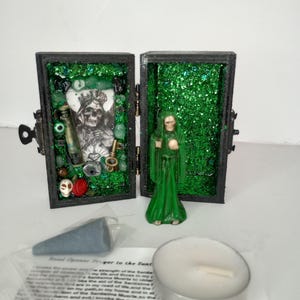 Puede incluir: Una peque&ntilde;a caja de madera con un interior de purpurina verde y una figurita de Santa Muerte. La caja est&aacute; abierta y contiene varios art&iacute;culos, incluyendo una calavera, cuentas y una botella. Tambi&eacute;n se incluye un cono de incienso azul verdoso y una vela de t&eacute; blanca.