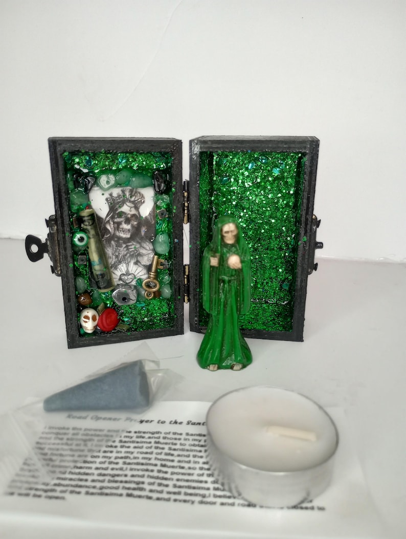 Puede incluir: Una peque&ntilde;a caja de madera con un interior de purpurina verde y una figurita de Santa Muerte. La caja est&aacute; abierta y contiene varios art&iacute;culos, incluyendo una calavera, cuentas y una botella. Tambi&eacute;n se incluye un cono de incienso azul verdoso y una vela de t&eacute; blanca.