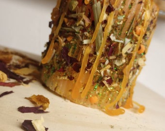 May include: A golden candle with a variety of dried flowers and herbs embedded in the wax. The candle has a drippy, melted wax design and is covered in glitter.