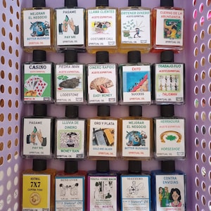 May include: A collection of small, rectangular glass bottles filled with colorful liquids, each labeled with Spanish text and illustrations. The bottles are arranged in a purple plastic basket. The labels feature phrases like "Pay Me" and "Better Business."