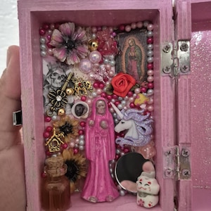 May include: A pink wooden box with an open lid, filled with various decorative elements. Inside are a miniature statue of Santa Muerte, a unicorn, a small bottle, and a picture of the Virgin Mary. The box is adorned with beads, flowers, and the text "Santa Muerte".