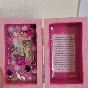 May include: A small pink wooden box with a hinged lid. The left side contains a collage of a hummingbird charm, a pink star, a key, beads, and a small image labeled Aphrodite. The right side features a printed prayer to Aphrodite under a glossy finish.