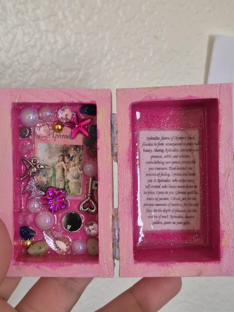 May include: A small pink wooden box with a hinged lid. The left side contains a collage of a hummingbird charm, a pink star, a key, beads, and a small image labeled Aphrodite. The right side features a printed prayer to Aphrodite under a glossy finish.