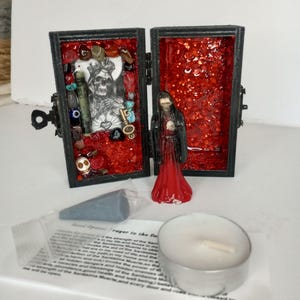 Puede incluir: Una peque&ntilde;a caja de madera negra con un interior rojo, que contiene una peque&ntilde;a figura de una persona con una t&uacute;nica roja, y una variedad de amuletos y baratijas. La caja est&aacute; abierta y la figura es visible. La caja est&aacute; etiquetada "Road Opener Prayer to the Santisima Muerte".