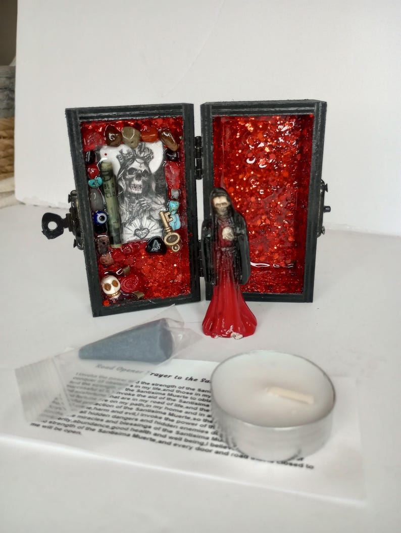 Puede incluir: Una peque&ntilde;a caja de madera negra con un interior rojo, que contiene una peque&ntilde;a figura de una persona con una t&uacute;nica roja, y una variedad de amuletos y baratijas. La caja est&aacute; abierta y la figura es visible. La caja est&aacute; etiquetada "Road Opener Prayer to the Santisima Muerte".