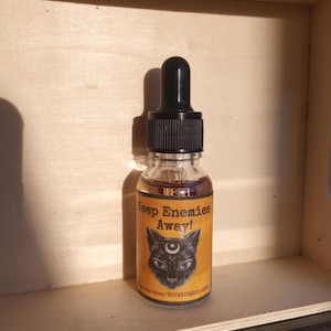 May include: A small glass bottle with a black dropper top and a yellow label that reads "Keep Enemies Away!" with a black cat illustration. The bottle is sitting on a wooden surface.