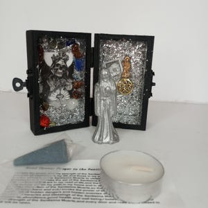 Puede incluir: Una peque&ntilde;a caja de madera negra con un interior de purpurina plateada, que contiene una figura plateada de una mujer y una calavera plateada con acentos rojos y azules. La caja est&aacute; abierta y se puede ver una peque&ntilde;a vela blanca y un cono de incienso azul en primer plano. El texto "Road Opener Prayer to the Santa Muerte" est&aacute; impreso en un papel blanco.