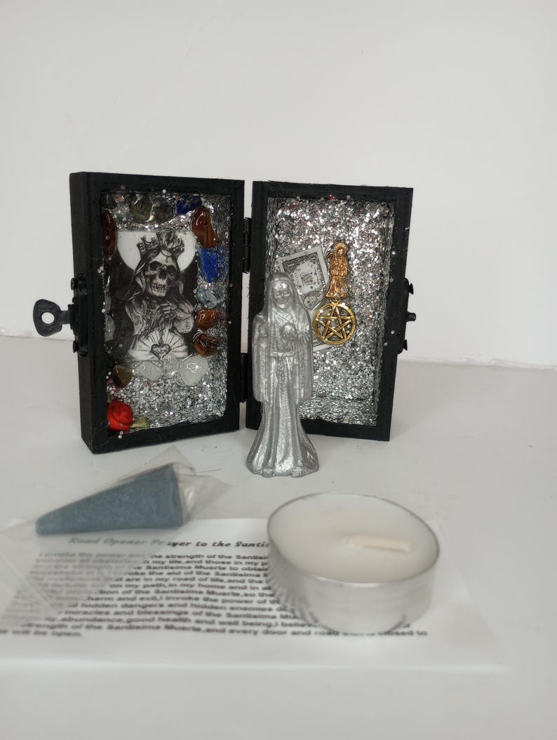 Puede incluir: Una peque&ntilde;a caja de madera negra con un interior de purpurina plateada, que contiene una figura plateada de una mujer y una calavera plateada con acentos rojos y azules. La caja est&aacute; abierta y se puede ver una peque&ntilde;a vela blanca y un cono de incienso azul en primer plano. El texto "Road Opener Prayer to the Santa Muerte" est&aacute; impreso en un papel blanco.