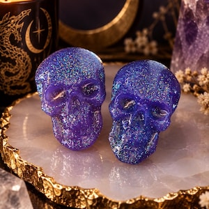 May include: Two small, three-dimensional skull figurines crafted from a translucent, deep purple resin material. Each skull is embedded with fine, multi-colored holographic glitter that catches the light, creating a shimmering, reflective surface effect.