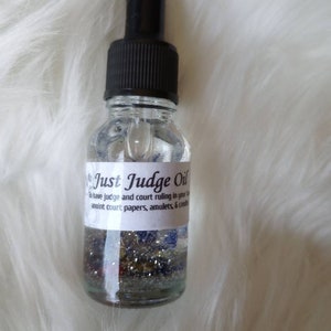 May include: A clear glass bottle with a black dropper top filled with a silver glitter liquid. The bottle has a white label that reads "Just Judge Oil" and includes instructions to anoint court papers, amulets, and candles.