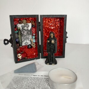 Puede incluir: Un peque&ntilde;o santuario de madera con un interior rojo, que presenta una figura esquel&eacute;tica y varios amuletos. La caja est&aacute; abierta, revelando su contenido. Una peque&ntilde;a figura de Santa Muerte se encuentra junto a la caja. Tambi&eacute;n se incluye una vela de t&eacute; blanca y un peque&ntilde;o cono de incienso gris.