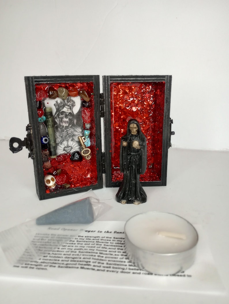 Puede incluir: Un peque&ntilde;o santuario de madera con un interior rojo, que presenta una figura esquel&eacute;tica y varios amuletos. La caja est&aacute; abierta, revelando su contenido. Una peque&ntilde;a figura de Santa Muerte se encuentra junto a la caja. Tambi&eacute;n se incluye una vela de t&eacute; blanca y un peque&ntilde;o cono de incienso gris.