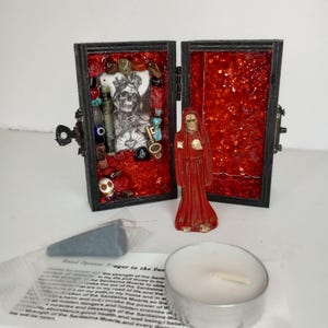 Puede incluir: Un peque&ntilde;o cofre de madera con un interior de purpurina roja. El cofre est&aacute; abierto y revela una peque&ntilde;a figura de Santa Muerte, una figura esquel&eacute;tica con una t&uacute;nica roja. El otro lado del cofre contiene una variedad de amuletos y baratijas, incluyendo una calavera, una llave y un coraz&oacute;n. El cofre est&aacute; acompa&ntilde;ado de una peque&ntilde;a vela blanca y un trozo de papel con una oraci&oacute;n a Santa Muerte.