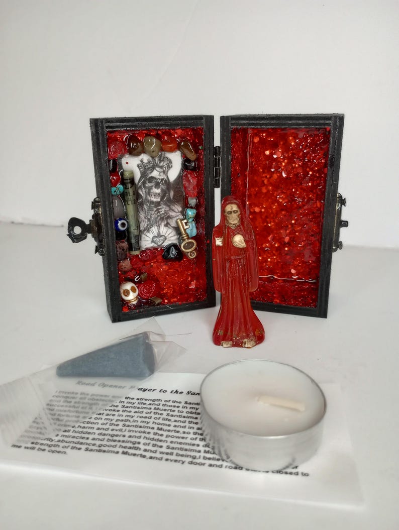 Puede incluir: Un peque&ntilde;o cofre de madera con un interior de purpurina roja. El cofre est&aacute; abierto y revela una peque&ntilde;a figura de Santa Muerte, una figura esquel&eacute;tica con una t&uacute;nica roja. El otro lado del cofre contiene una variedad de amuletos y baratijas, incluyendo una calavera, una llave y un coraz&oacute;n. El cofre est&aacute; acompa&ntilde;ado de una peque&ntilde;a vela blanca y un trozo de papel con una oraci&oacute;n a Santa Muerte.
