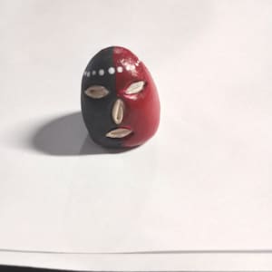 May include: A small, rounded object painted with a split black and red design. It features embedded white shell-like pieces forming eyes, a nose, and a mouth, with a horizontal row of small white dots painted across the upper forehead area.
