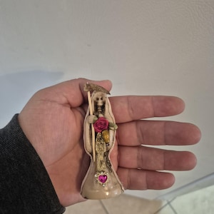 May include: A small, off-white figurine of a skeletal figure in a hooded robe, holding a scythe and roses. The figurine has a pink heart-shaped jewel on the chest. The figure is held in a hand.