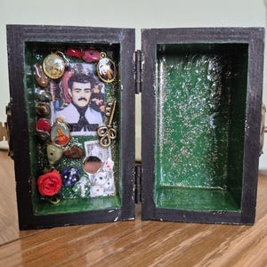 May include: A black wooden box with a hinged lid, featuring a green glitter interior. The left side contains a portrait of a man, religious charms, a small key, a red rose, dice, and a miniature dollar bill. The right side is empty and green.