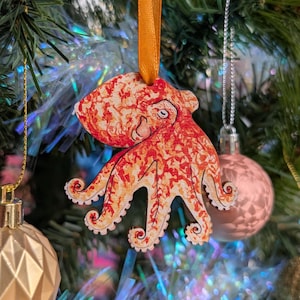 May include: A wooden octopus Christmas decoration with a red and orange pattern, suspended by an orange ribbon. The ornament is on a Christmas tree with other decorations and tinsel.