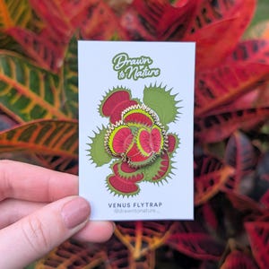 Könnte beinhalten: Eine weiße Karte mit einem Venusfliegenfalle-Anstecker. Der Anstecker hat ein rot-grünes Design mit dem Text "Drawn to Nature" und "Venus Flytrap". Der Hintergrund ist eine Darstellung von rotem und grünem Laub.