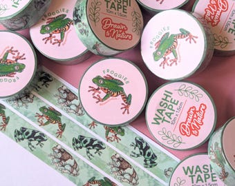 May include: A collection of washi tape rolls and a strip of washi tape featuring frog illustrations. The rolls have pink and green accents with the words "Washi Tape" and "Froggies" printed on them. The tape is 10m x 1.5cm.