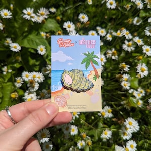 May include: A green and brown enamel pin of a seashell on a beach with a palm tree in the background. The pin says "Drawn to Nature x Heather Filby" at the top.