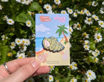 May include: A green and brown enamel pin of a seashell on a beach with a palm tree in the background. The pin says "Drawn to Nature x Heather Filby" at the top.