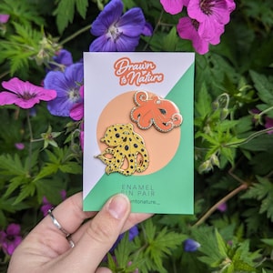 May include: A pair of enamel pins featuring two different octopus designs. The octopus on the left is yellow with black spots and the octopus on the right is orange. The pins are on a card that says "Drawn to Nature" and "Enamel Pin Pair".