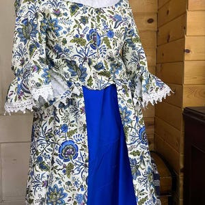 May include: A historical costume featuring a white and blue floral patterned jacket with bell sleeves and a white lace collar. The costume includes a long, royal blue skirt. The sleeves and collar are trimmed with white lace.