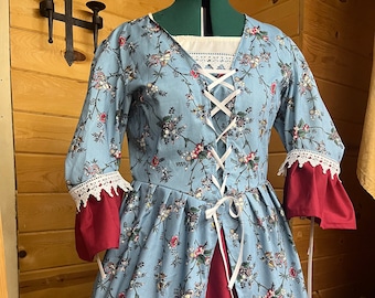 May include: A blue and white floral print dress with a red under layer. The dress has a lace trim on the sleeves and a white lace neckline. The dress is laced up the front with a white ribbon.