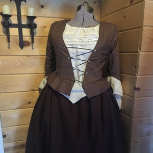 以下が含まれることがあります： マネキンに着せられた茶色とクリーム色の歴史的衣装。衣装は、長い濃い茶色のスカート、レーストリム付きのクリーム色のブラウス、ひもで締めたフロントの茶色のジャケットが特徴です。袖にはクリーム色の袖口が付いています。