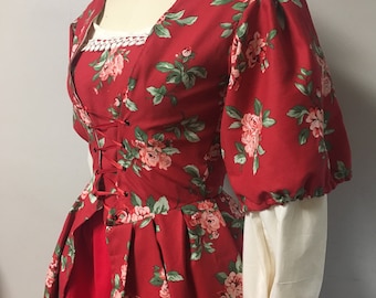May include: A red dress with a floral print and a white lace trim at the neckline. The dress has a fitted bodice with a lace-up front and a full skirt. The sleeves are puffed and end in a white cuff.