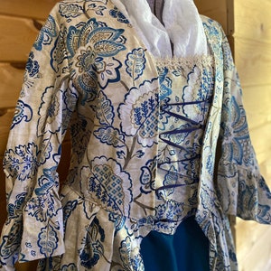 Może przedstawiać: Kremowa kurtka z niebieskim i brązowym wzorem kwiatowym. Kurtka ma koronkową lamówkę przy dekolcie i niebieskie sznurowanie z przodu. Kurtka jest noszona na turkusowej spódnicy.