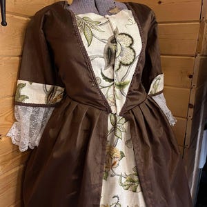May include: A brown historical dress with a floral pattern on the bodice and sleeves. The dress features a full skirt and lace trim on the sleeves. The bodice has a cream-colored background with green and yellow floral designs.