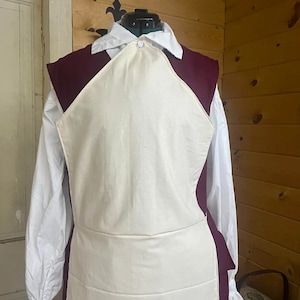 May include: A white apron with a burgundy vest, worn over a white shirt with ruffled cuffs.