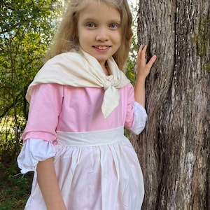 May include: A young girl wearing a pink dress with a white apron and a white scarf tied around her neck. She is standing next to a tree and smiling.
