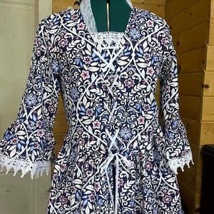 May include: A long-sleeved dress with a floral pattern in navy, white, light blue, and pink. The dress features a white lace collar and cuffs, and a white ribbon corset detail. The dress is displayed on a dress form.