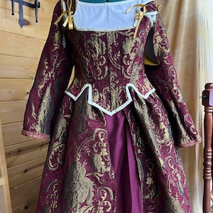 May include: A burgundy and gold brocade dress with a white lace collar and gold ribbon accents. The dress has a fitted bodice and a full skirt.