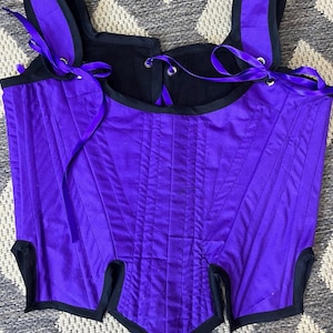 May include: Purple corset with black trim and ribbon ties. The corset has a pointed bottom and a fitted bodice.