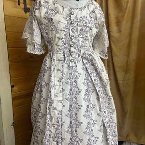 May include: A white dress with a floral pattern in purple. The dress has a lace trim on the sleeves and a row of buttons down the front.