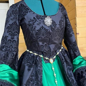 May include: A black and green dress with a damask pattern. The dress has a white pearl necklace and a matching belt with a gold buckle and a small pendant. The dress has long sleeves with green satin cuffs and black lace trim.